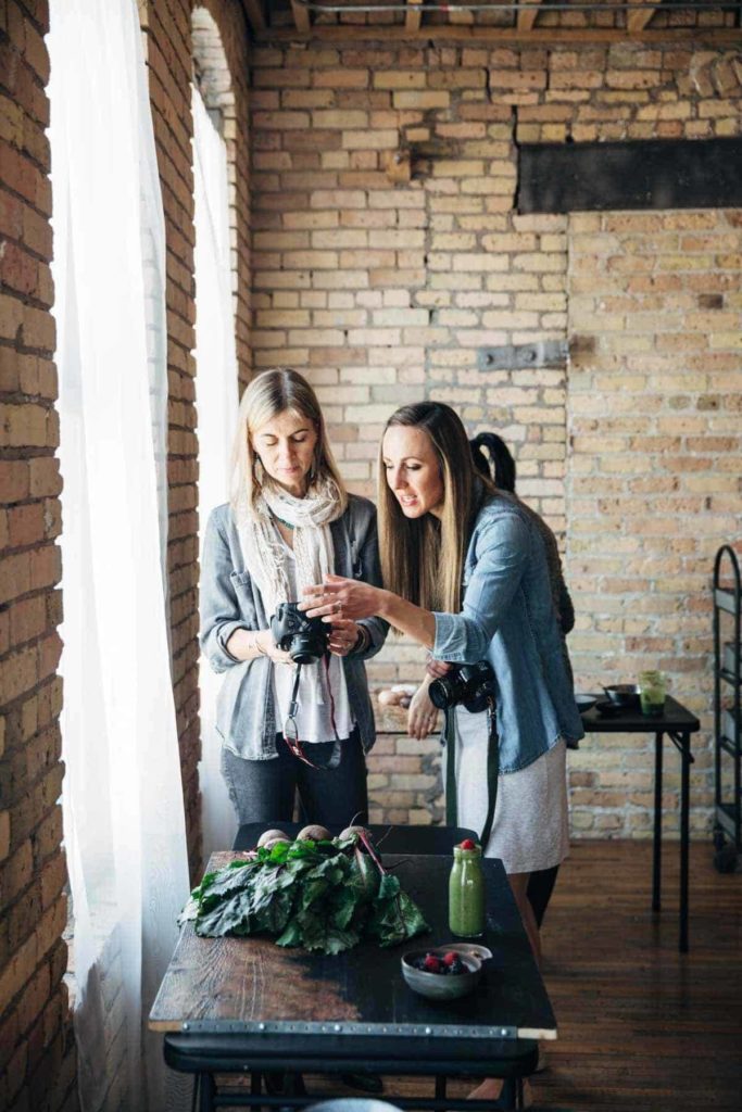 Two women working with DSLR cameras to photograph a table of food.