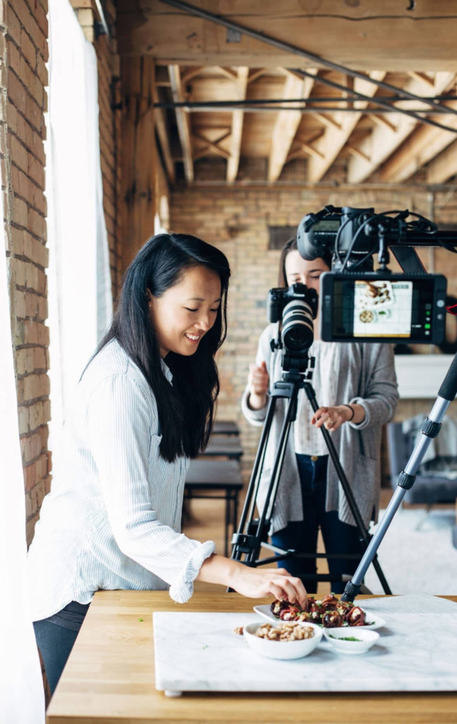 One woman arranging food on a plate and another shooting video from a camera on a tripod
